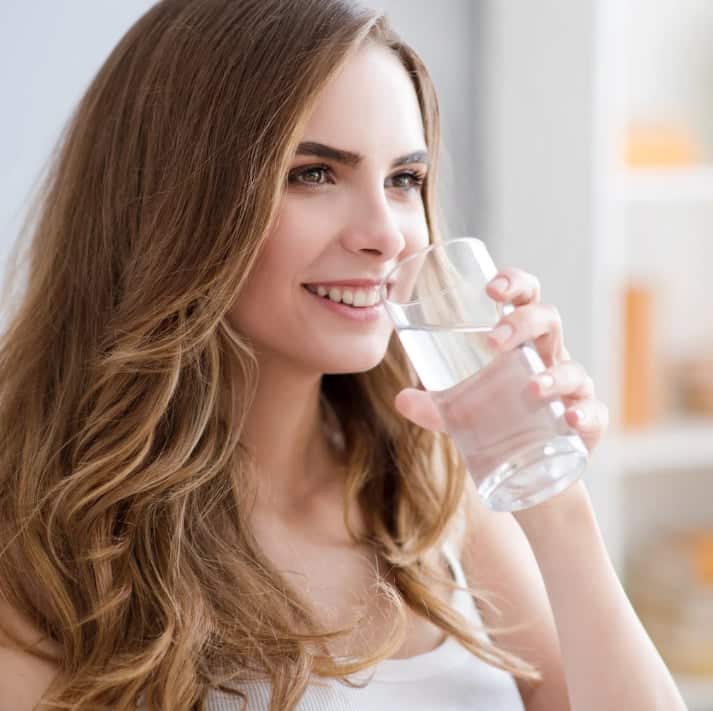 Woman with long hair smiling and drinking a glass of water to stay hydrated and protect her oral health.